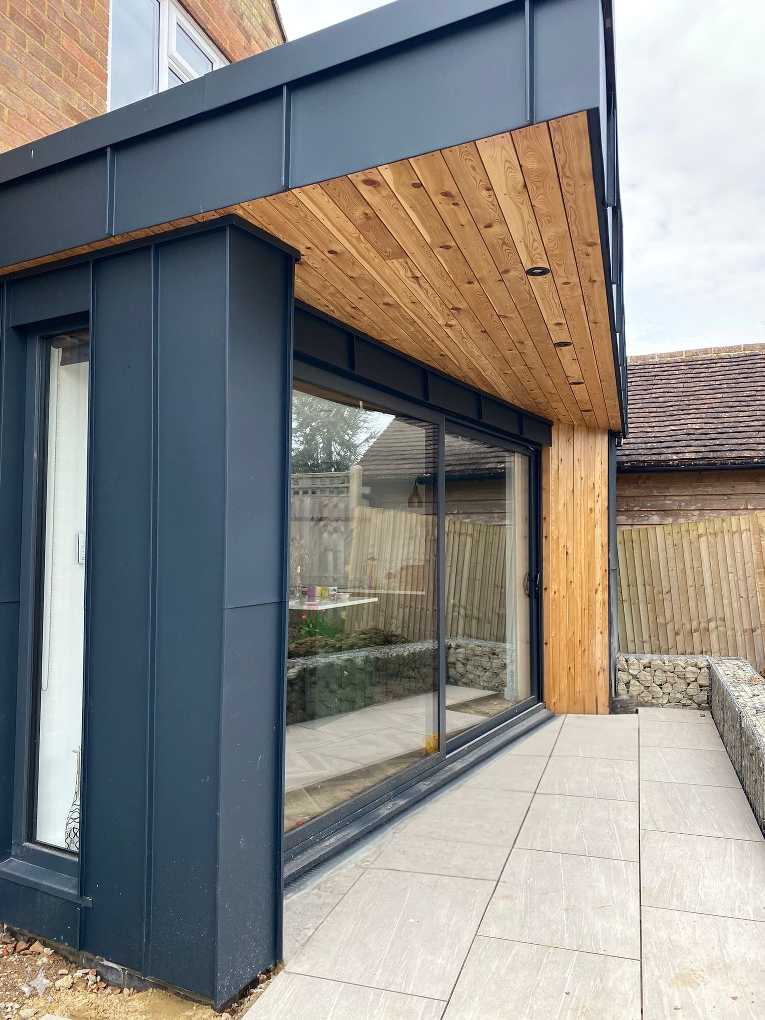 Modern architectural extension with black metal cladding and wood accents. Sliding glass doors open to a stone-tiled patio. Fenced yard with gabion baskets filled with rocks visible in the background. The roof overhang features wooden paneling.